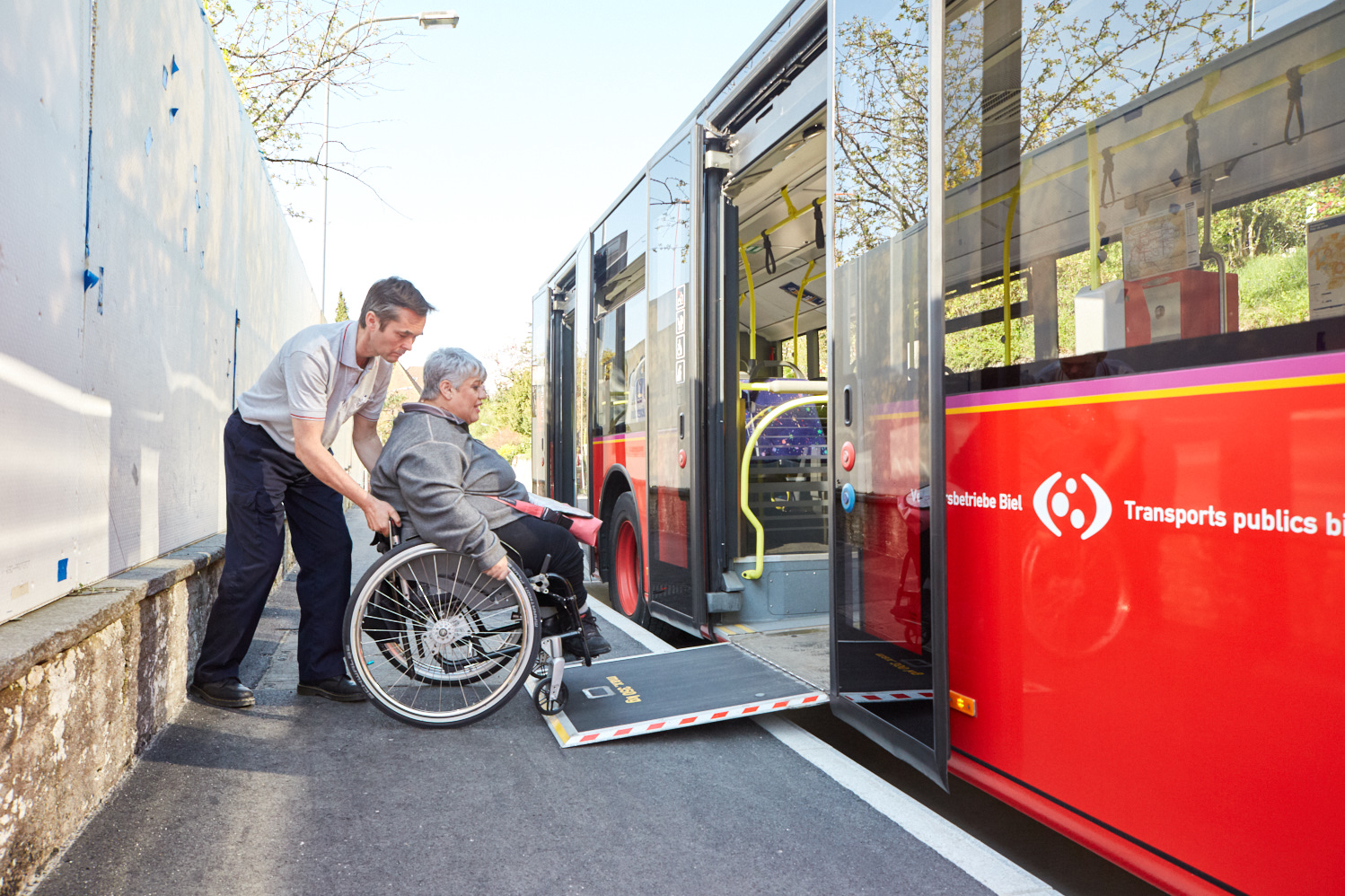 Accessibilité: Une personne en fauteuil roulant est aidée par un accompagnant pour monter dans un bus rouge via une rampe déployée.