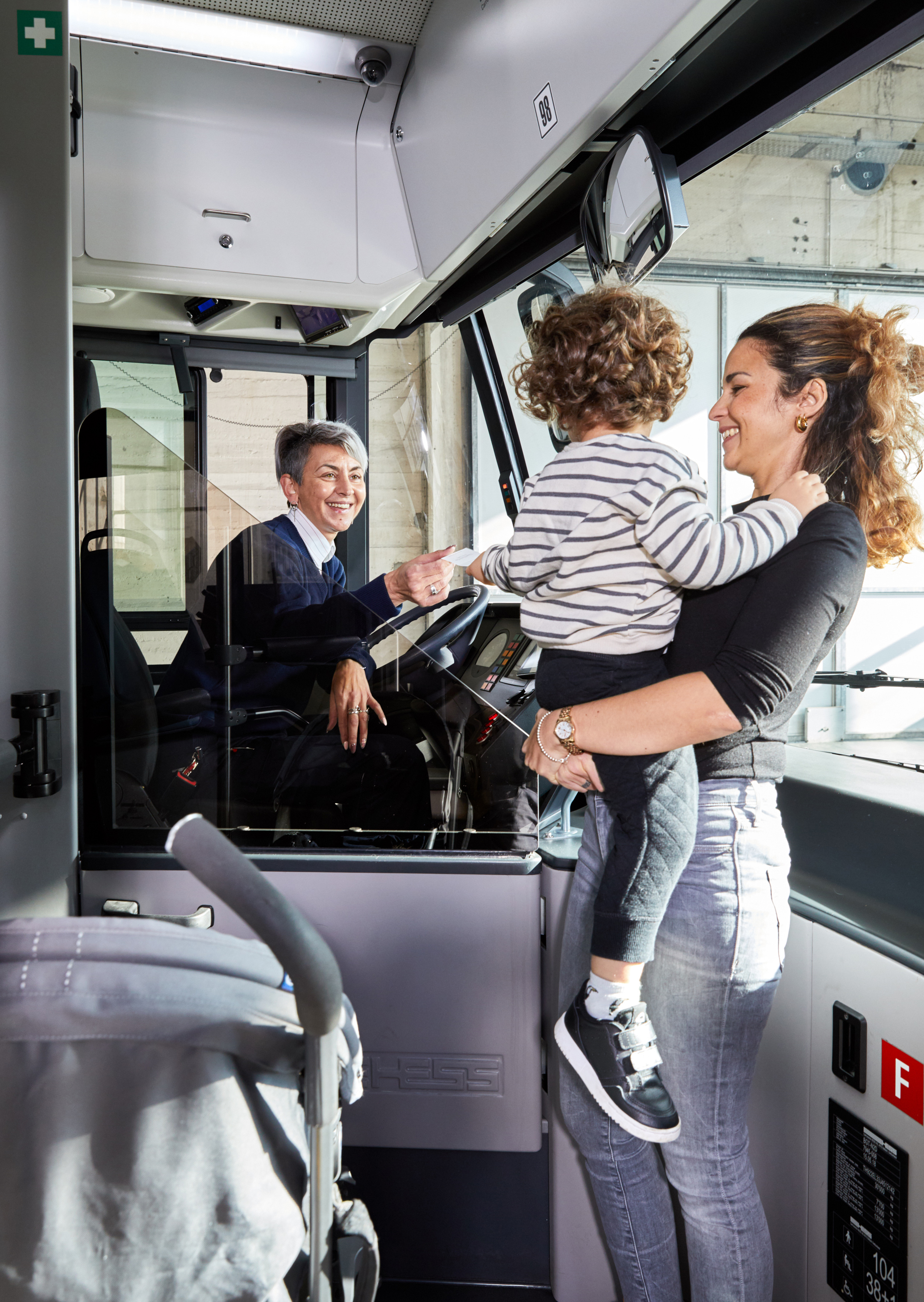 Notre_manière_d’être_et_d’agir: Une personne adulte tenant un jeune enfant se tient à l’entrée d’un bus et interagit avec la personne au poste de conduite.